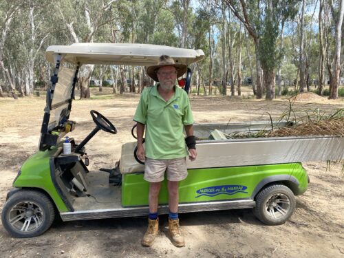 George Dick, a man with a short beard and glasses. He is wearing his work uniform of a green polo shirt, shorts, work boots and a wide brimmed hat standing proudly in front of his green work golf kart in the holiday park gardens.