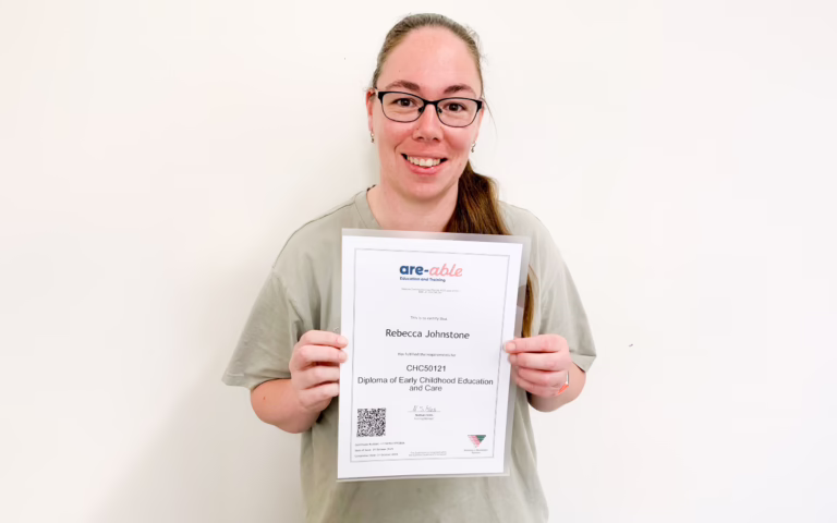 A person wearing glasses smiles at the camera while holding up a framed certificate against a plain light wall.