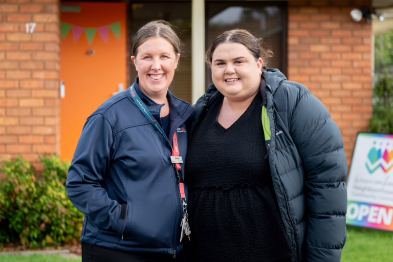 Two women standing close together and smiling at the camera outside a brick community building, with an orange door, bunting, and a signboard in the background. The woman on the left is an are-able team member wearing a navy jacket, name badge, and lanyard with keys. The woman on the right is wearing a black top and a dark puffer jacket.