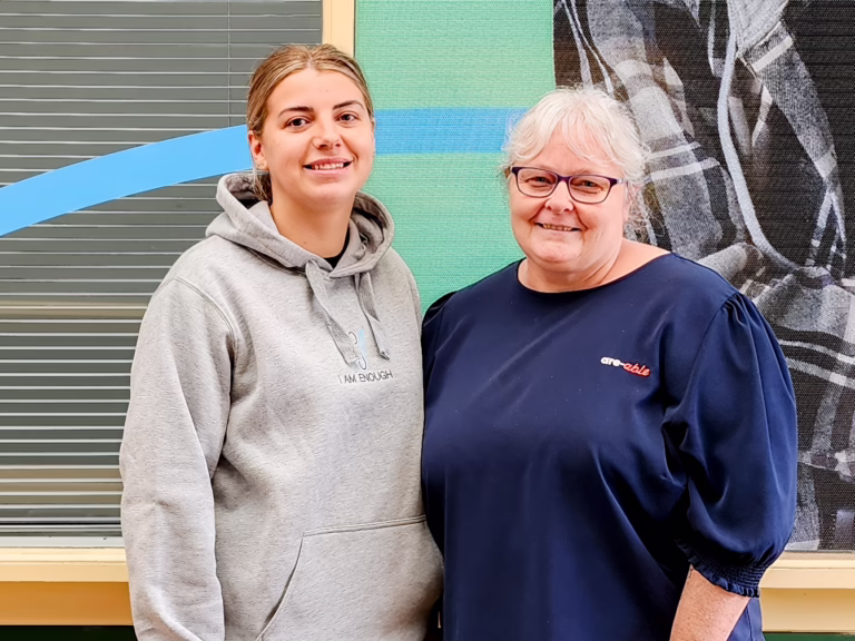 Two women stand side by side smiling at the camera. One wears a grey hoodie, and the other is an are-able team member wearing a navy top with the are-able logo.