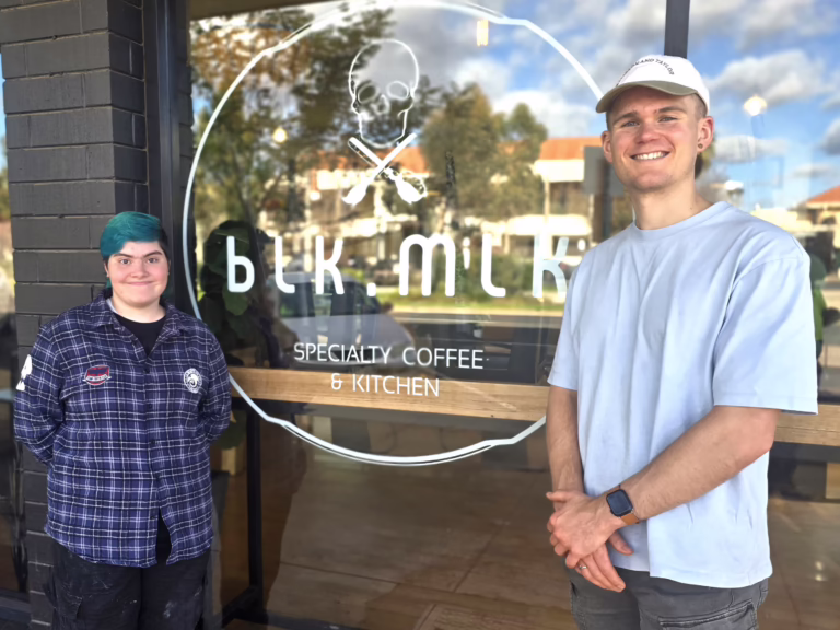 Two people stand outside a café window with a skull and crossed tools logo. One wears a checked shirt and has turquoise hair, and the other wears a light blue T-shirt and white cap.