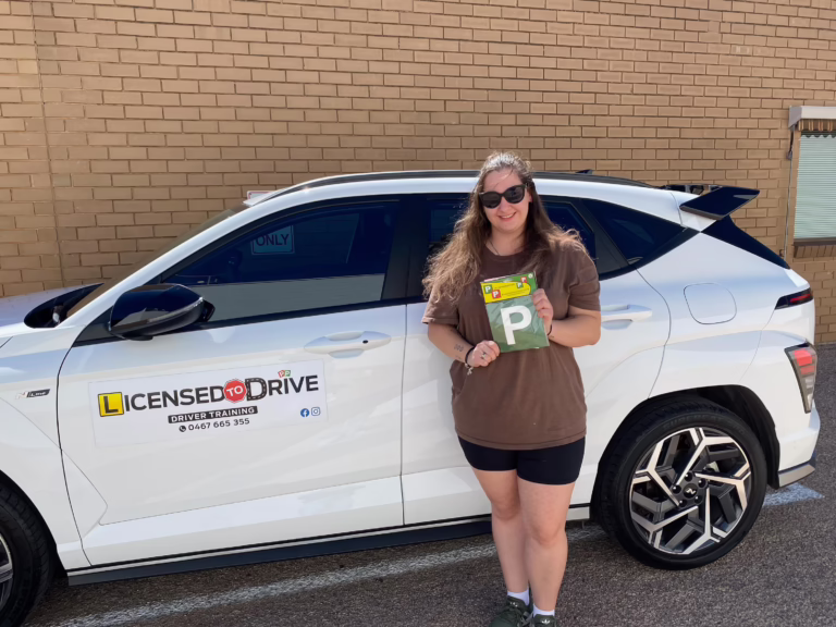 A smiling person wearing sunglasses stands beside a white car marked as a driver training vehicle, holding a green P plate.