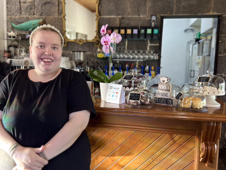 A smiling person in a black top and pearl-style headband stands beside a wooden café counter with glass-covered cakes, an orchid and coffee equipment behind.