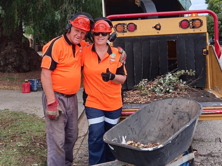 Two people in orange tree work clothing, helmets and ear protection stand beside a woodchipper loaded with branches and leaves. A wheelbarrow sits in the foreground.