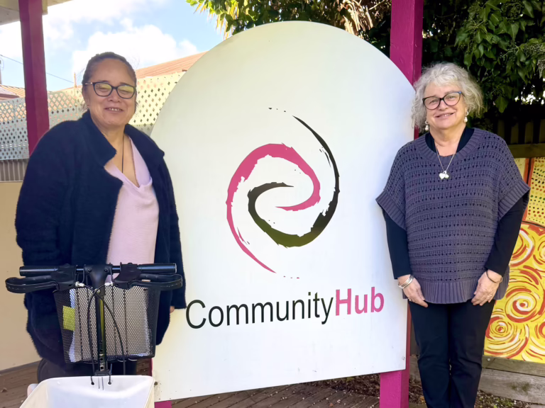 Two smiling women wearing glasses stand either side of a large CommunityHub sign outdoors, with a mobility scooter’s handlebars and basket in the foreground.